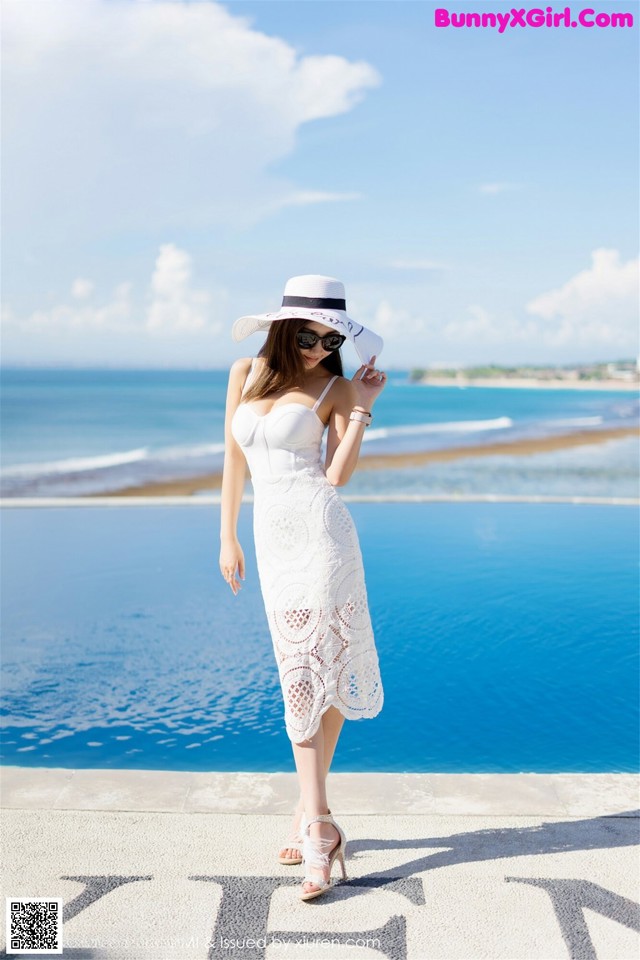 A woman in a white dress and hat standing by a pool.