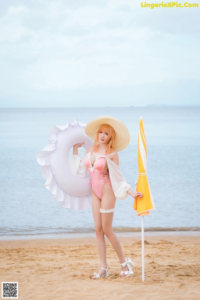 A woman in a pink bathing suit and hat holding an umbrella on the beach.